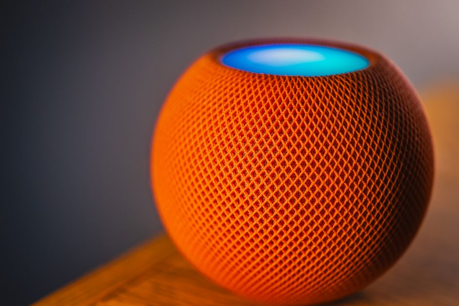 An orange spherical fabric smart speaker with a glowing top, on a wooden shelf